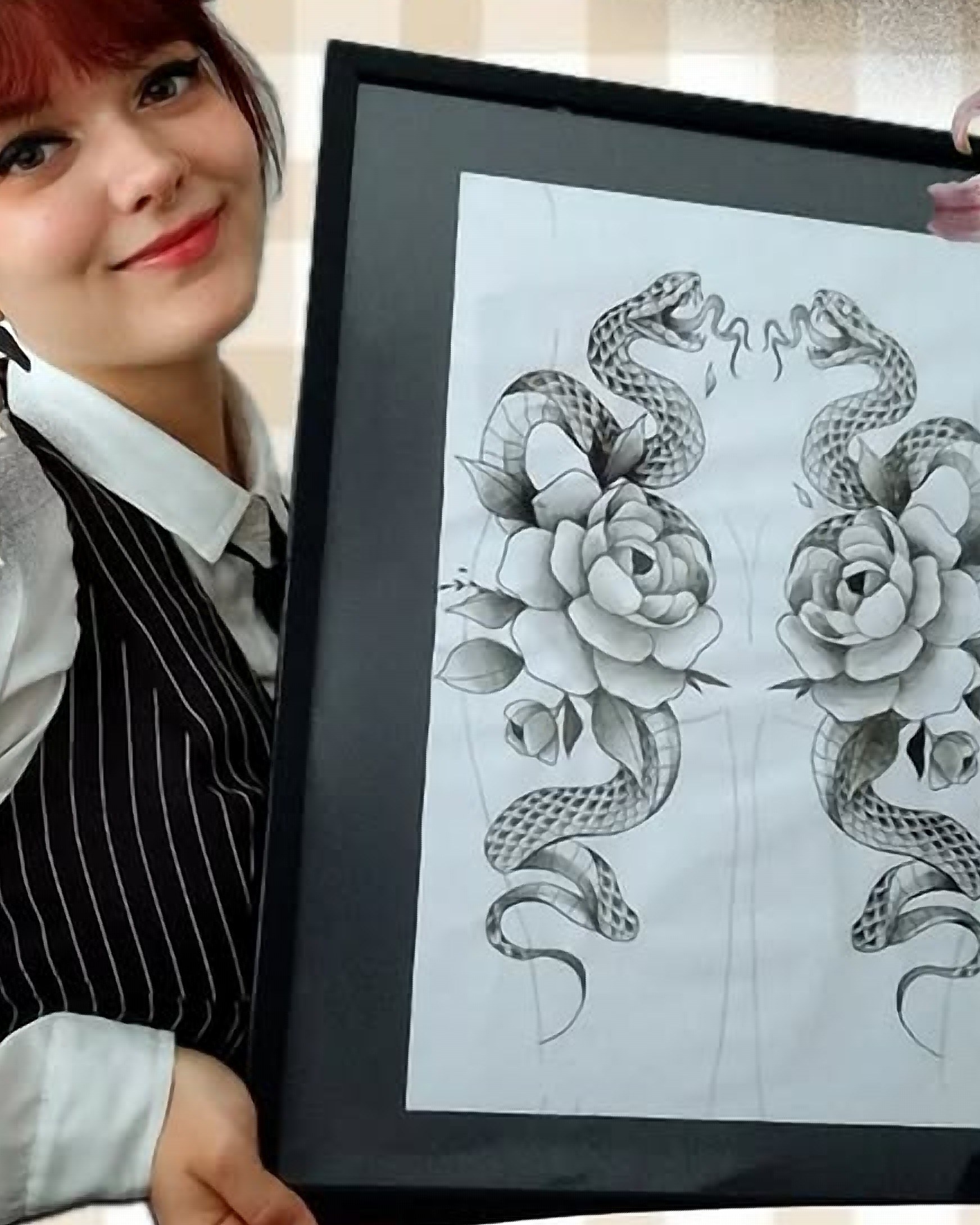Kael'ink souriante, en gilet rayé, tenant l'un de ses dessins encadrés : deux serpents entourés de roses au trait fin.
